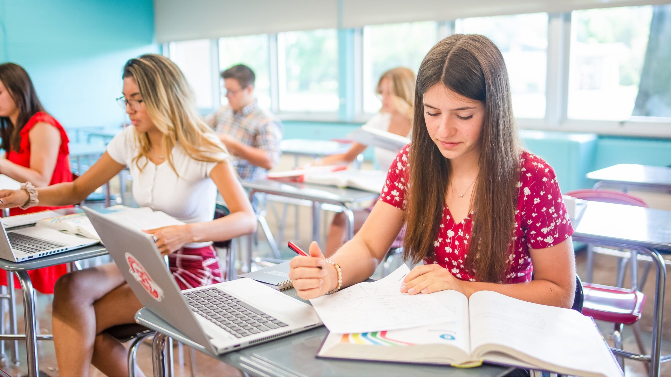 Girl student in classroom sitting at a desk with an open book and laptop holding a pencil